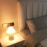 Table lamp with a plain cream pleated shade and a pale green ceramic base with a raised chevron pattern lit on a bedside cabinet, plugged into a wall socket.