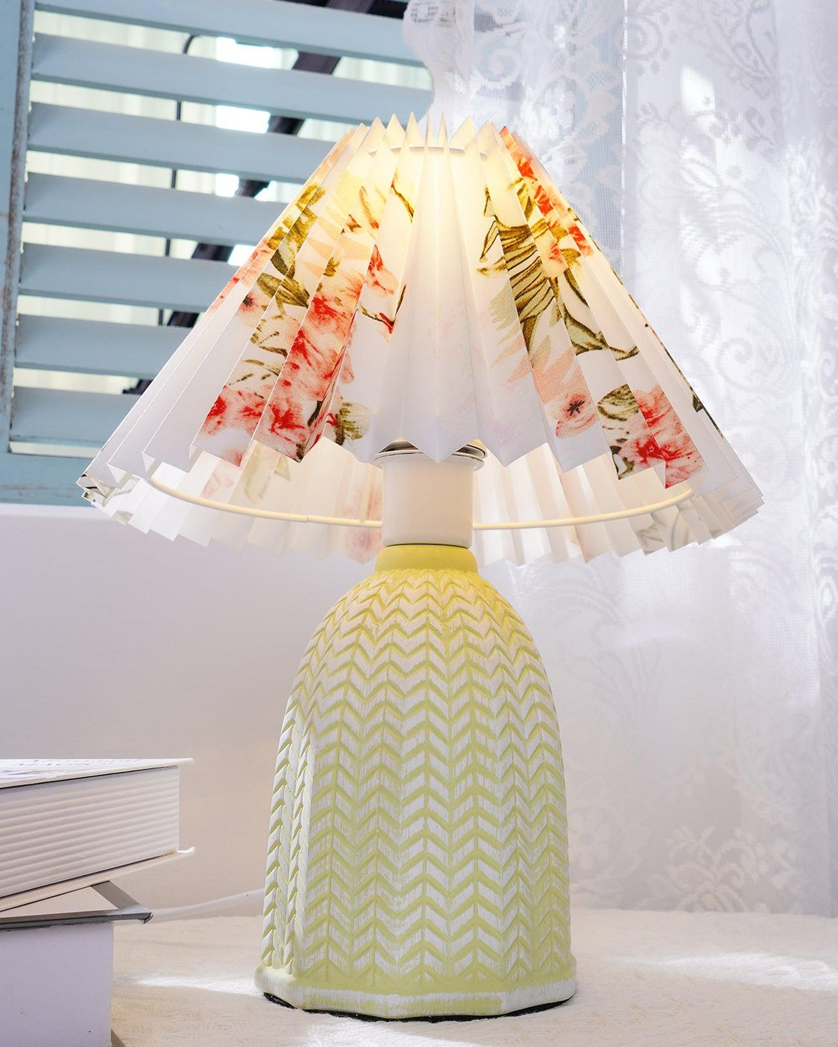 Table lamp with a white pleated shade in a printed floral pattern and a pale green ceramic base with a raised chevron pattern lit on a windowsill beside a book.