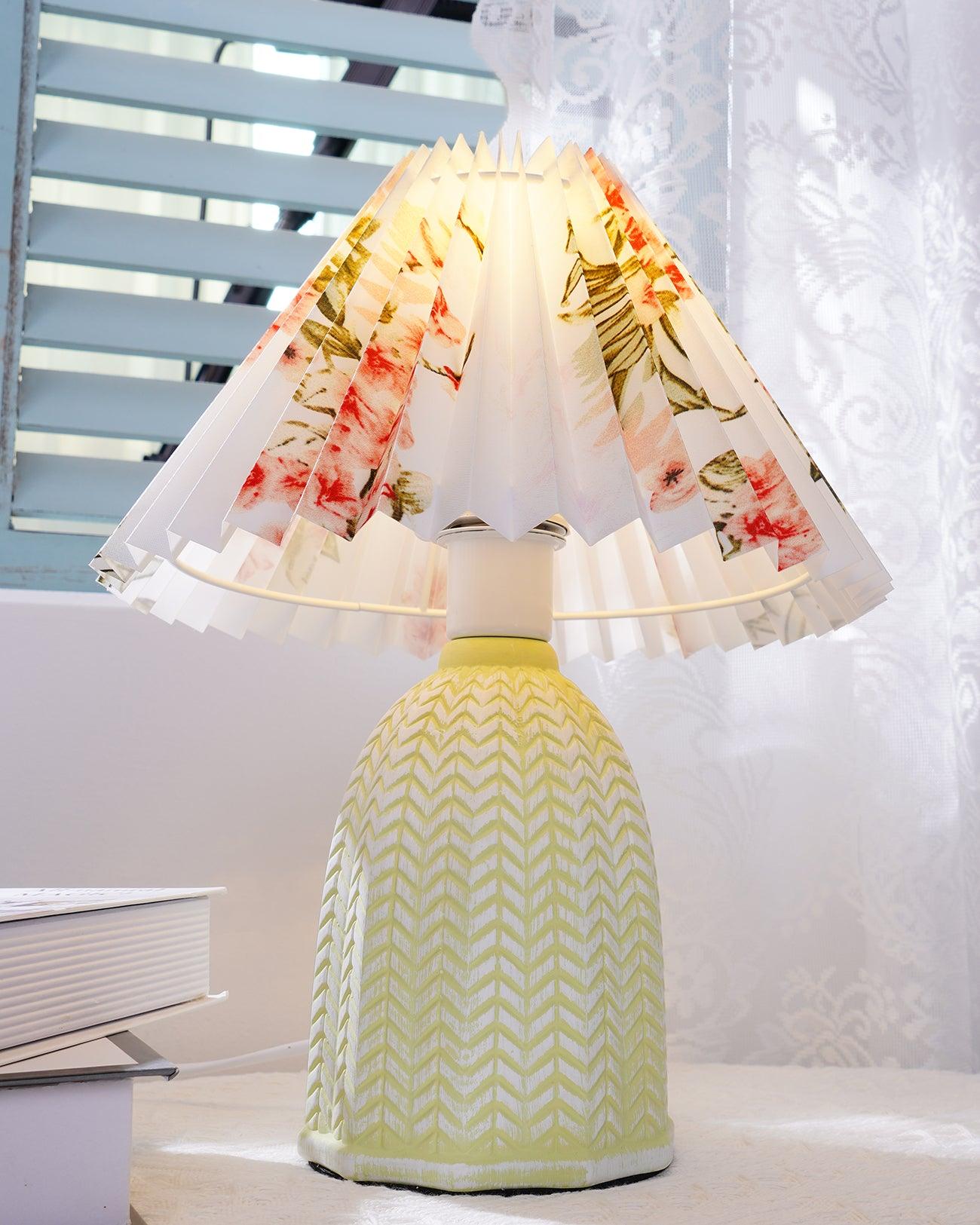 Table lamp with a white pleated shade in a printed floral pattern and a pale green ceramic base with a raised chevron pattern lit on a windowsill beside a book.