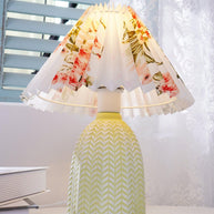 Table lamp with a white pleated shade in a printed floral pattern and a pale green ceramic base with a raised chevron pattern lit on a windowsill beside a book.