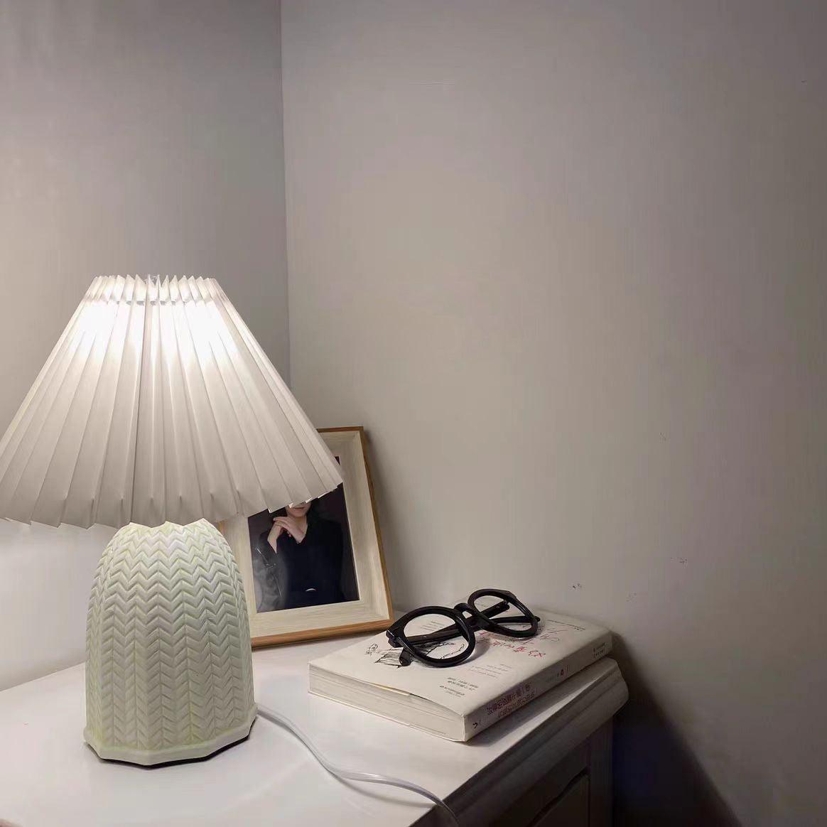 Table lamp with a plain cream pleated shade and a pale green ceramic base with a raised chevron pattern lit on a bedside table beside glasses and a book.