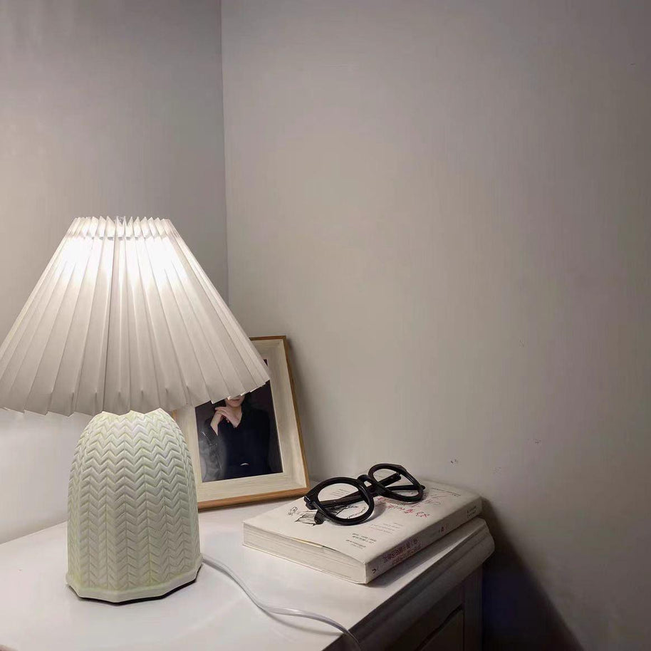 Table lamp with a plain cream pleated shade and a pale green ceramic base with a raised chevron pattern lit on a bedside table beside glasses and a book.