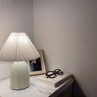 Table lamp with a plain cream pleated shade and a pale green ceramic base with a raised chevron pattern lit on a bedside table beside glasses and a book.