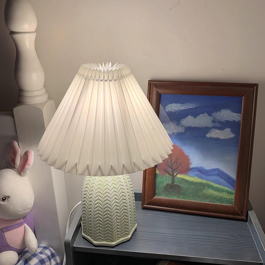 Table lamp with a pleated shade and a ridged pale green base lit on a bedside table beside a framed picture, seen a little closer.