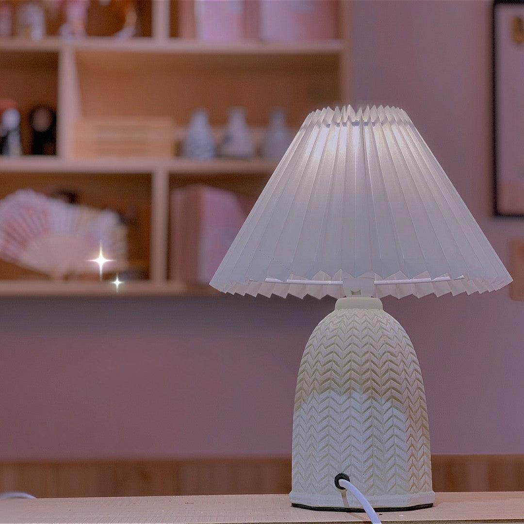 Table lamp with a plain cream pleated shade and a pale green ceramic base with a raised chevron pattern lit in front of a shelf of small objects.
