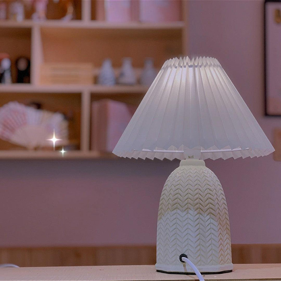 Table lamp with a plain cream pleated shade and a pale green ceramic base with a raised chevron pattern lit in front of a shelf of small objects.