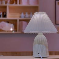 Table lamp with a plain cream pleated shade and a pale green ceramic base with a raised chevron pattern lit in front of a shelf of small objects.