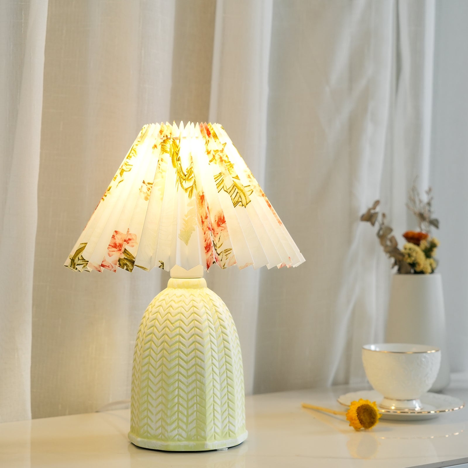 Table lamp with a white pleated shade in a printed floral pattern and a pale green ceramic base with a raised chevron pattern lit beside a curtain and a vase of dried flowers.