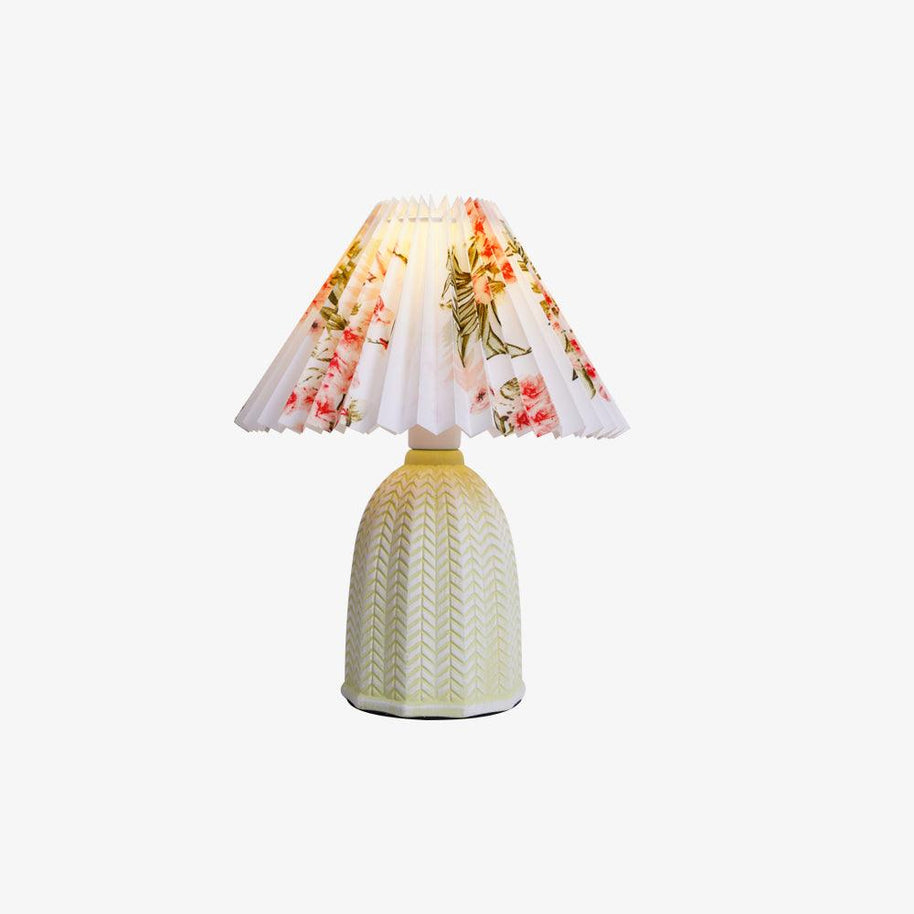 Table lamp with a white pleated shade in a printed floral pattern and a pale green ceramic base with a raised chevron pattern lit on a white background.