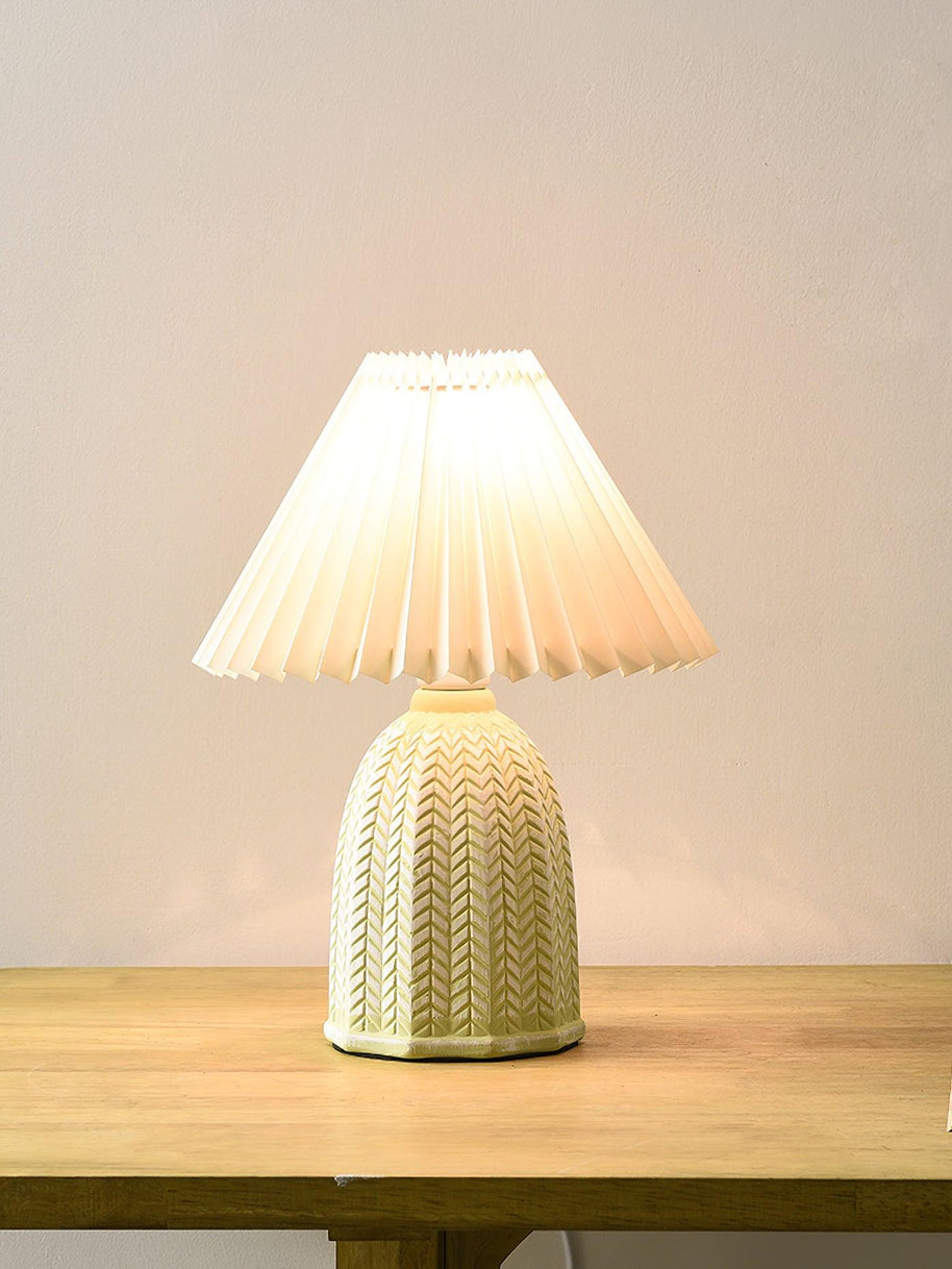 Table lamp with a plain cream pleated shade and a pale green ceramic base with a raised chevron pattern on a wooden table against a plain wall.