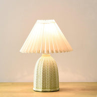 Table lamp with a plain cream pleated shade and a pale green ceramic base with a raised chevron pattern on a wooden table against a plain wall.