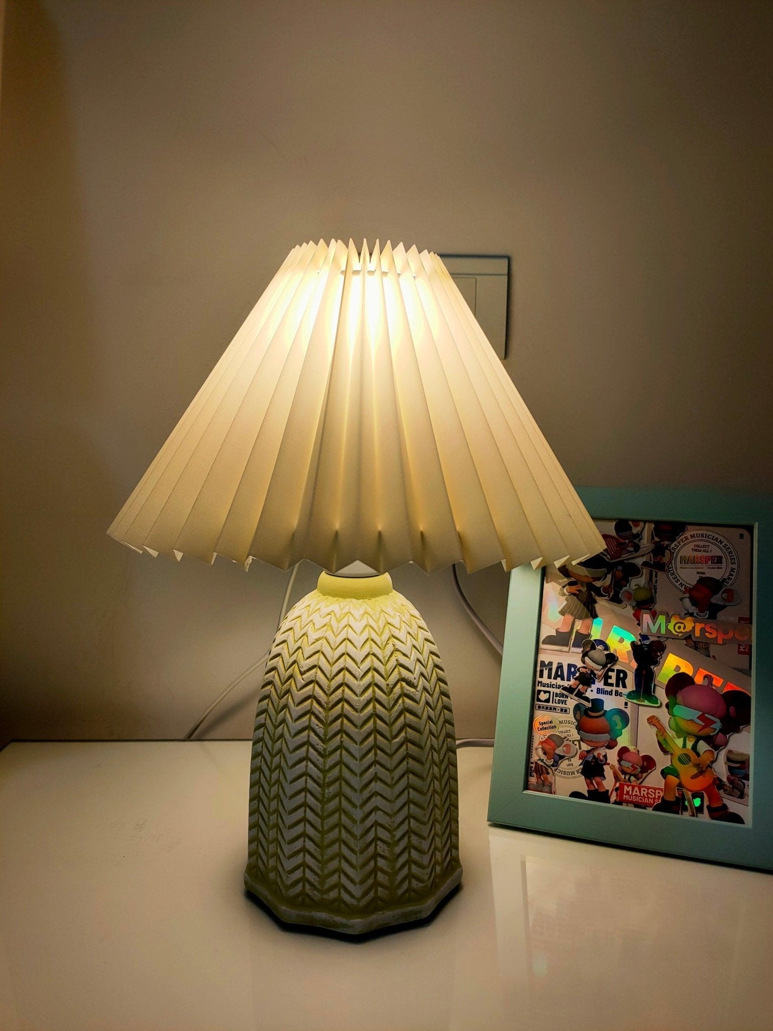 Table lamp with a plain cream pleated shade and a pale green ceramic base with a raised chevron pattern lit on a table beside a framed picture.