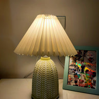Table lamp with a plain cream pleated shade and a pale green ceramic base with a raised chevron pattern lit on a table beside a framed picture.