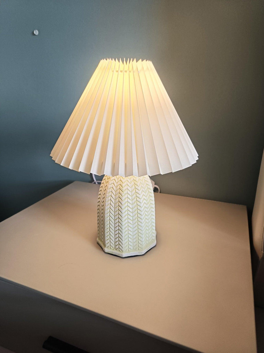 Table lamp with a plain cream pleated shade and a pale green ceramic base with a raised chevron pattern lit on a table against a dark blue wall.
