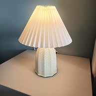 Table lamp with a plain cream pleated shade and a pale green ceramic base with a raised chevron pattern lit on a table against a dark blue wall.