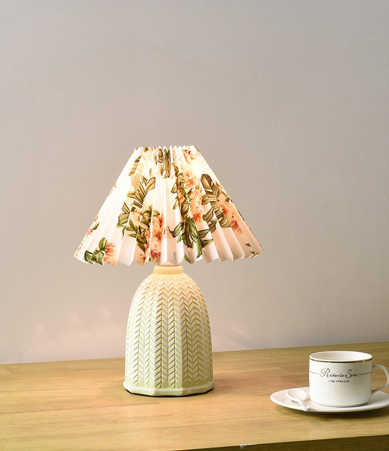 Table lamp with a white pleated shade in a printed floral pattern and a pale green ceramic base with a raised chevron pattern on a wooden table beside a teacup.