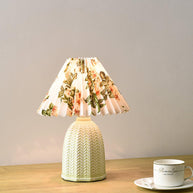 Table lamp with a white pleated shade in a printed floral pattern and a pale green ceramic base with a raised chevron pattern on a wooden table beside a teacup.