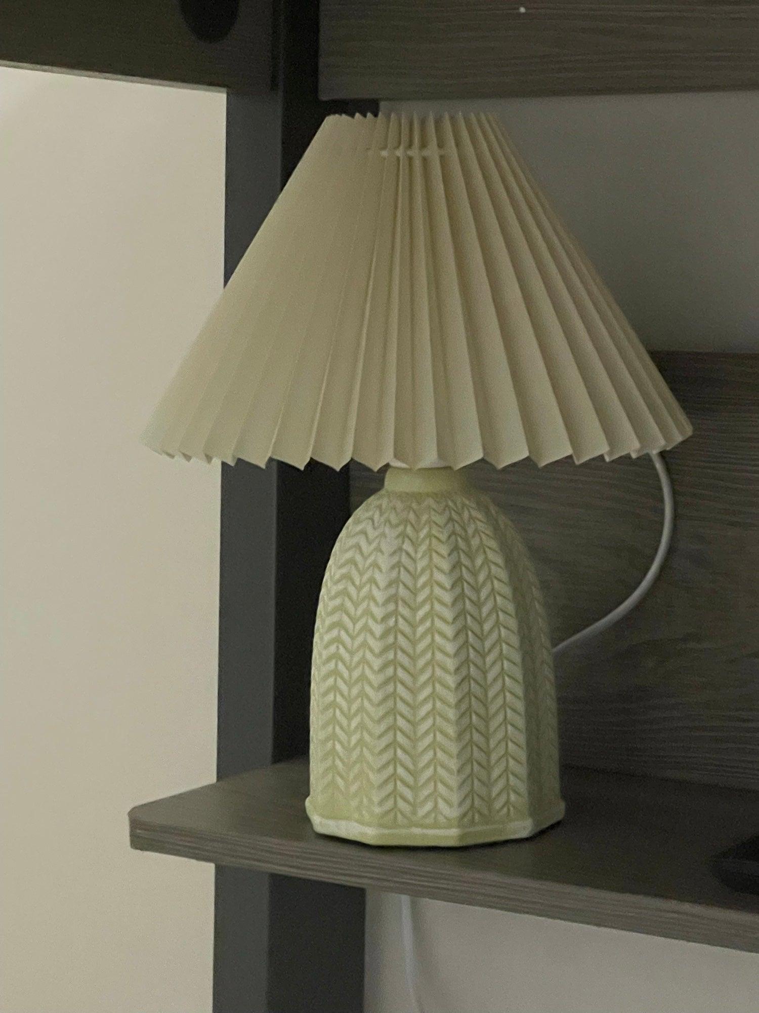 Table lamp with a plain cream pleated shade and a pale green ceramic base with a raised chevron pattern on a shelf.