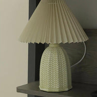 Table lamp with a plain cream pleated shade and a pale green ceramic base with a raised chevron pattern on a shelf.