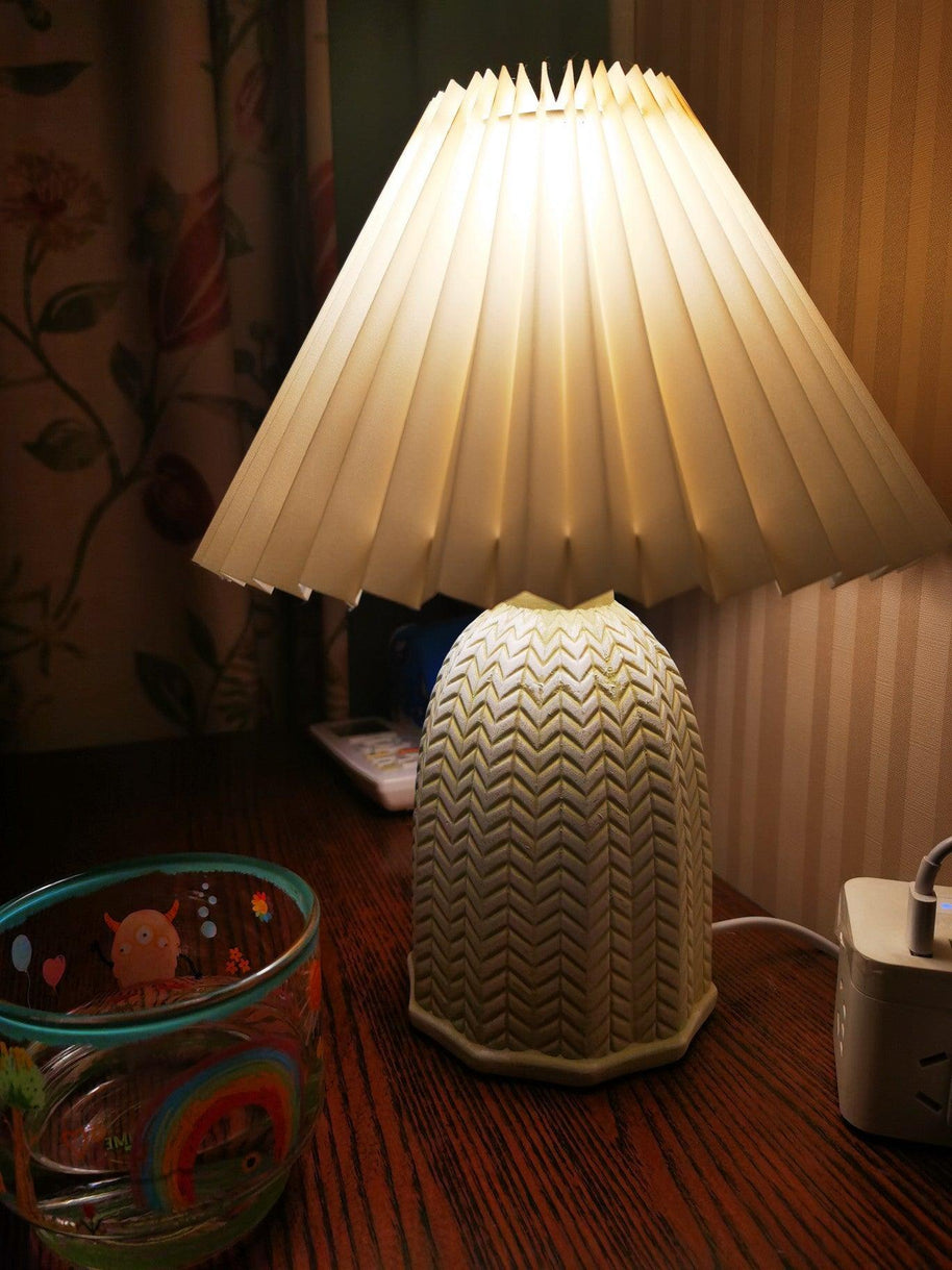 Table lamp with a plain cream pleated shade and a pale green ceramic base with a raised chevron pattern lit on a wooden surface beside a glass bowl.
