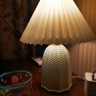 Table lamp with a plain cream pleated shade and a pale green ceramic base with a raised chevron pattern lit on a wooden surface beside a glass bowl.
