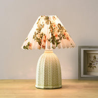 Table lamp with a white pleated shade in a printed floral pattern and a pale green ceramic base with a raised chevron pattern lit on a wooden table beside a framed picture.