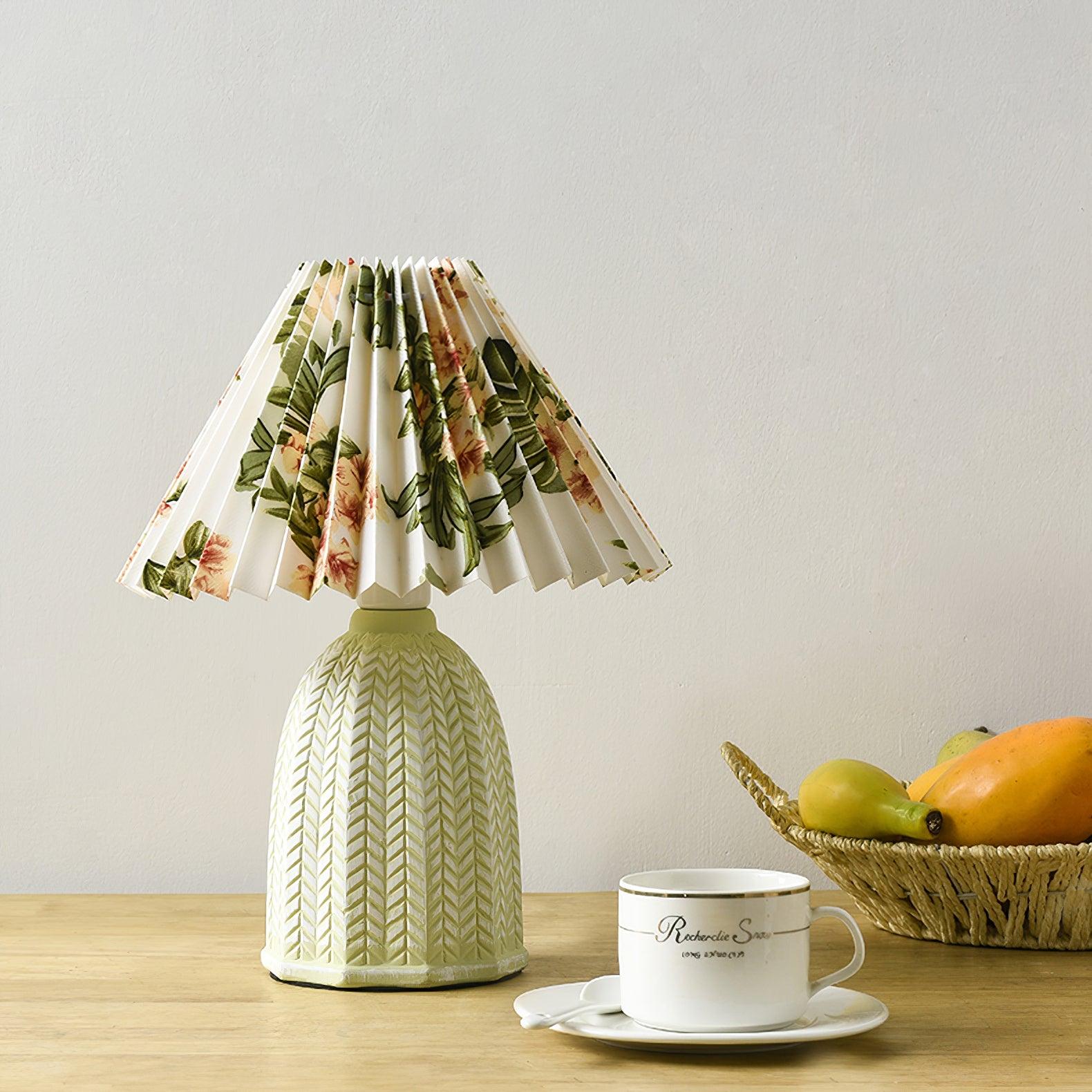 Table lamp with a white pleated shade in a printed floral pattern and a pale green ceramic base with a raised chevron pattern lit on a wooden table beside a fruit basket.