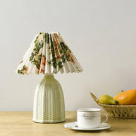 Table lamp with a white pleated shade in a printed floral pattern and a pale green ceramic base with a raised chevron pattern lit on a wooden table beside a fruit basket.