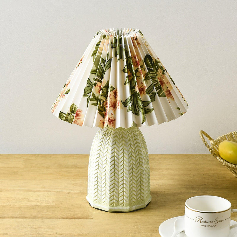 Table lamp with a white pleated shade in a printed floral pattern and a pale green ceramic base with a raised chevron pattern lit on a wooden table beside a teacup.