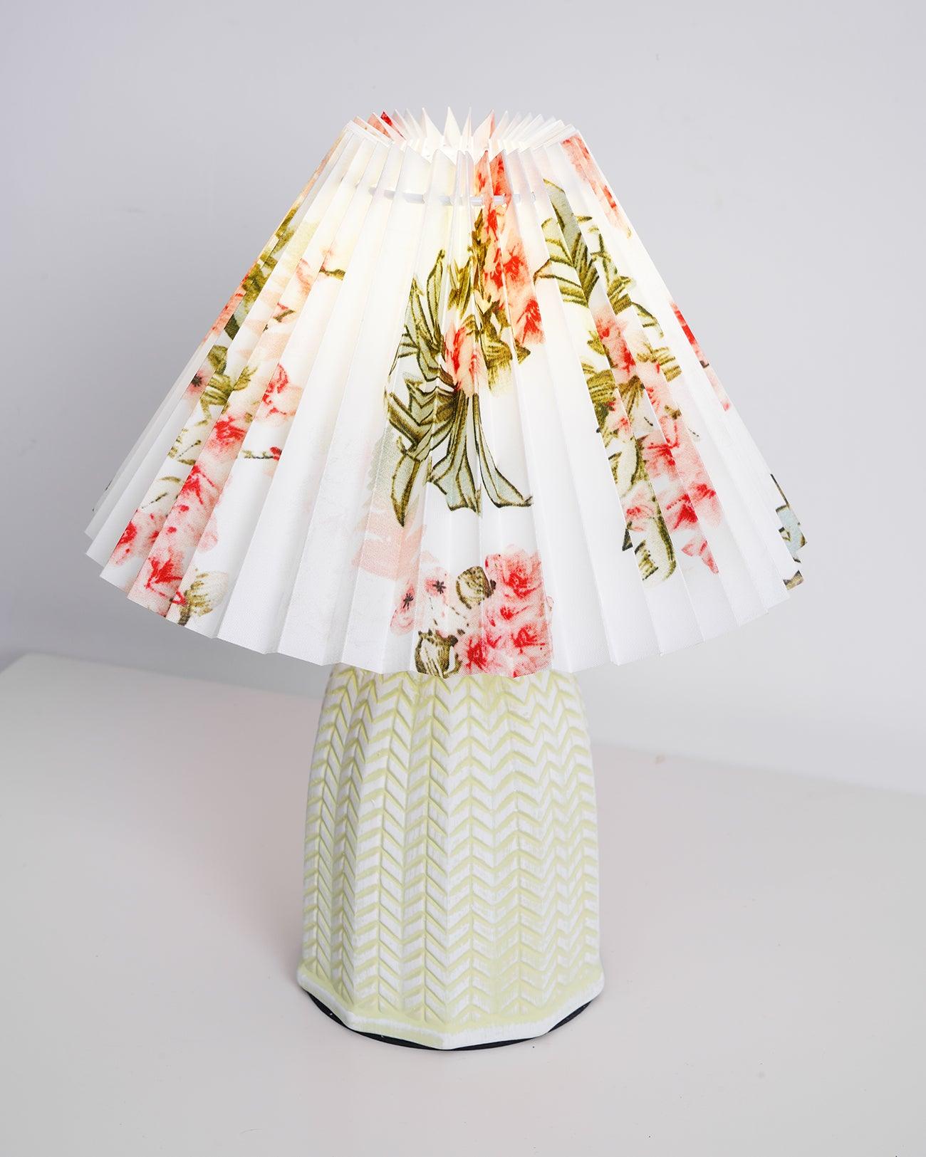 Table lamp with a white pleated shade in a printed floral pattern and a pale green ceramic base with a raised chevron pattern, shown on a pale background with a wide shade.