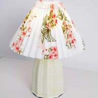 Table lamp with a white pleated shade in a printed floral pattern and a pale green ceramic base with a raised chevron pattern, shown on a pale background with a wide shade.