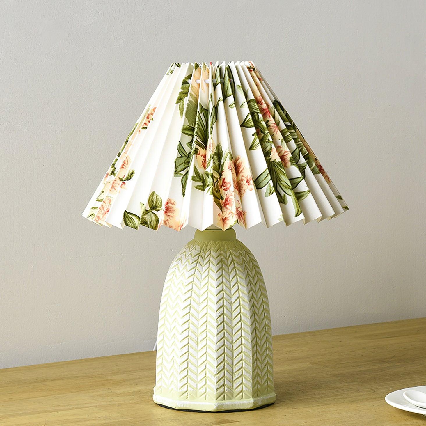 Table lamp with a white pleated shade in a printed floral pattern and a pale green ceramic base with a raised chevron pattern lit on a wooden table.