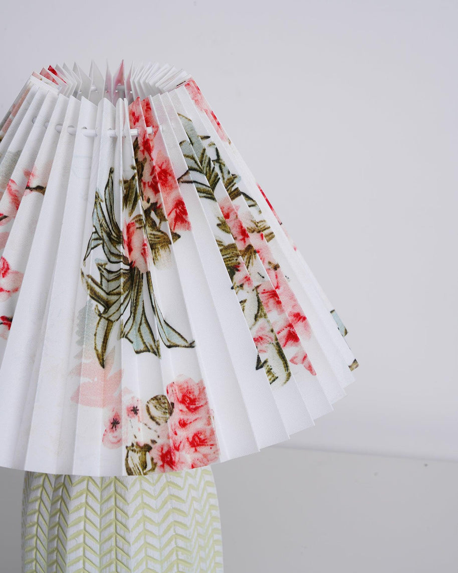 Table lamp with a white pleated shade in a printed floral pattern and a pale green ceramic base with a raised chevron pattern, seen close from below on a plain background.