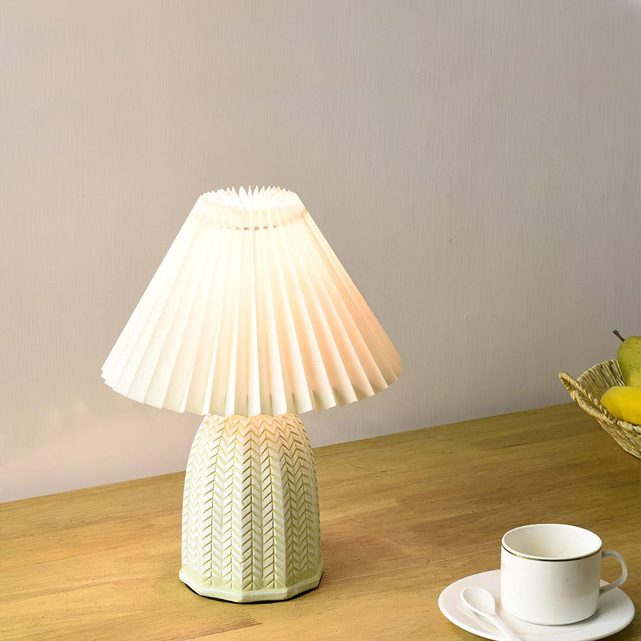 Table lamp with a plain cream pleated shade and a pale green ceramic base with a raised chevron pattern lit on a wooden table beside a cup.