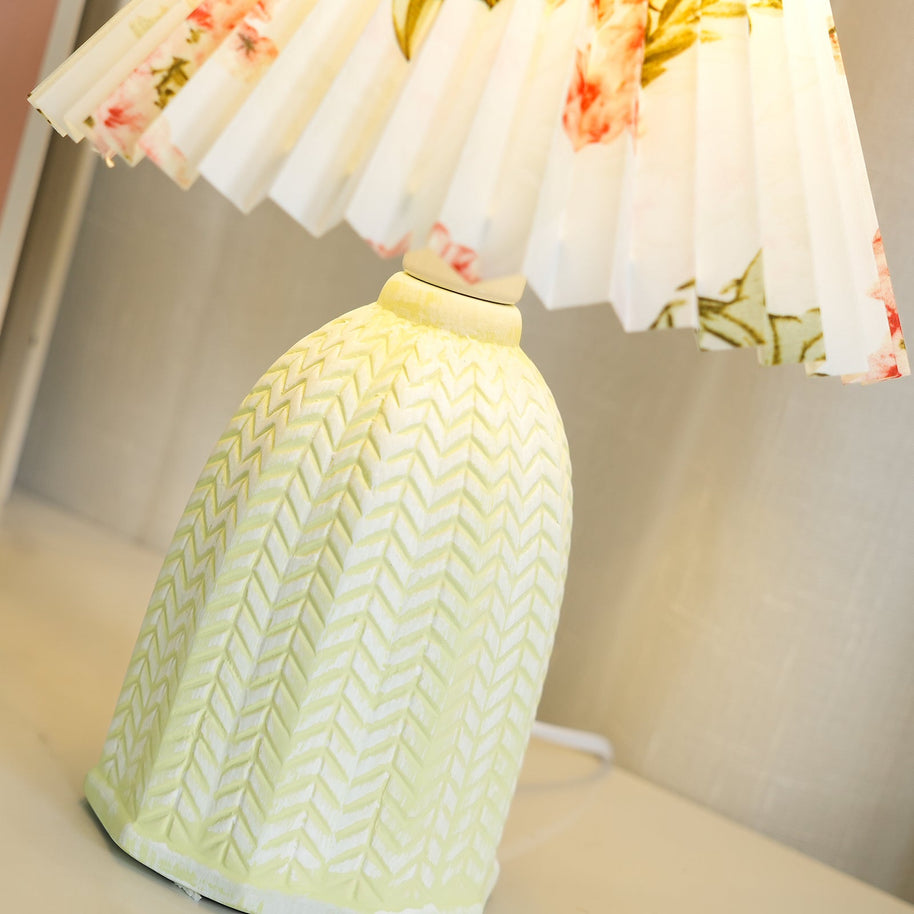 Table lamp with a white pleated shade in a printed floral pattern and a pale green ceramic base with a raised chevron pattern lit, seen close beside the base.