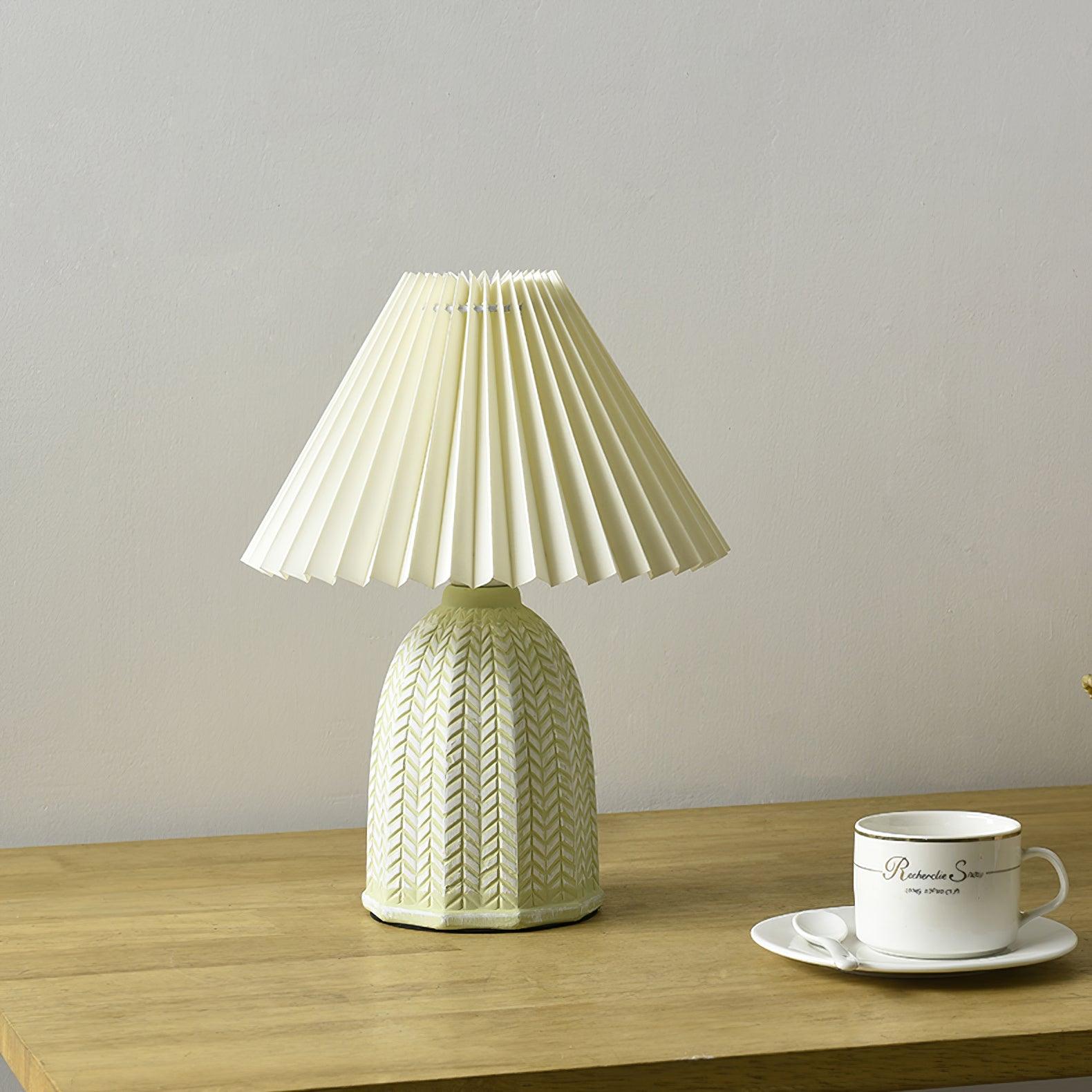 Table lamp with a plain cream pleated shade and a pale green ceramic base with a raised chevron pattern on a wooden table beside a teacup.