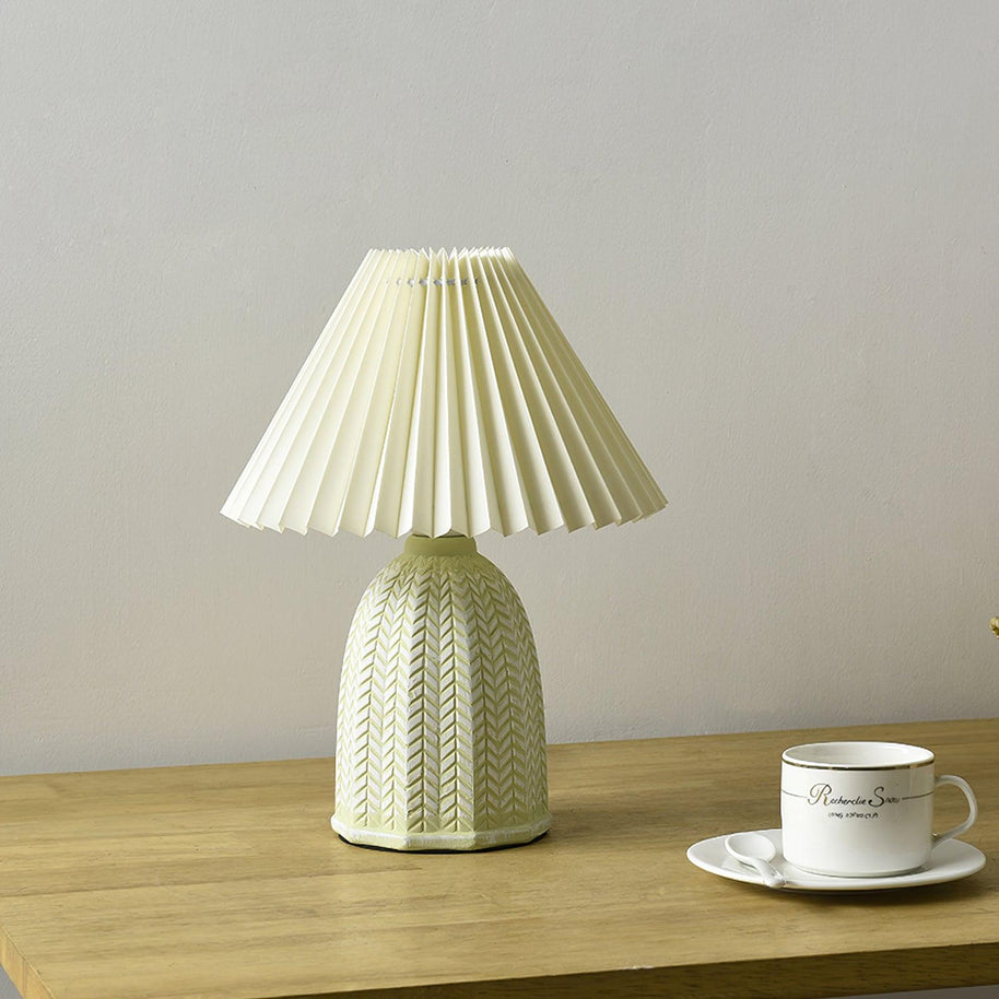 Table lamp with a plain cream pleated shade and a pale green ceramic base with a raised chevron pattern on a wooden table beside a teacup.