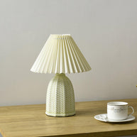 Table lamp with a plain cream pleated shade and a pale green ceramic base with a raised chevron pattern on a wooden table beside a teacup.
