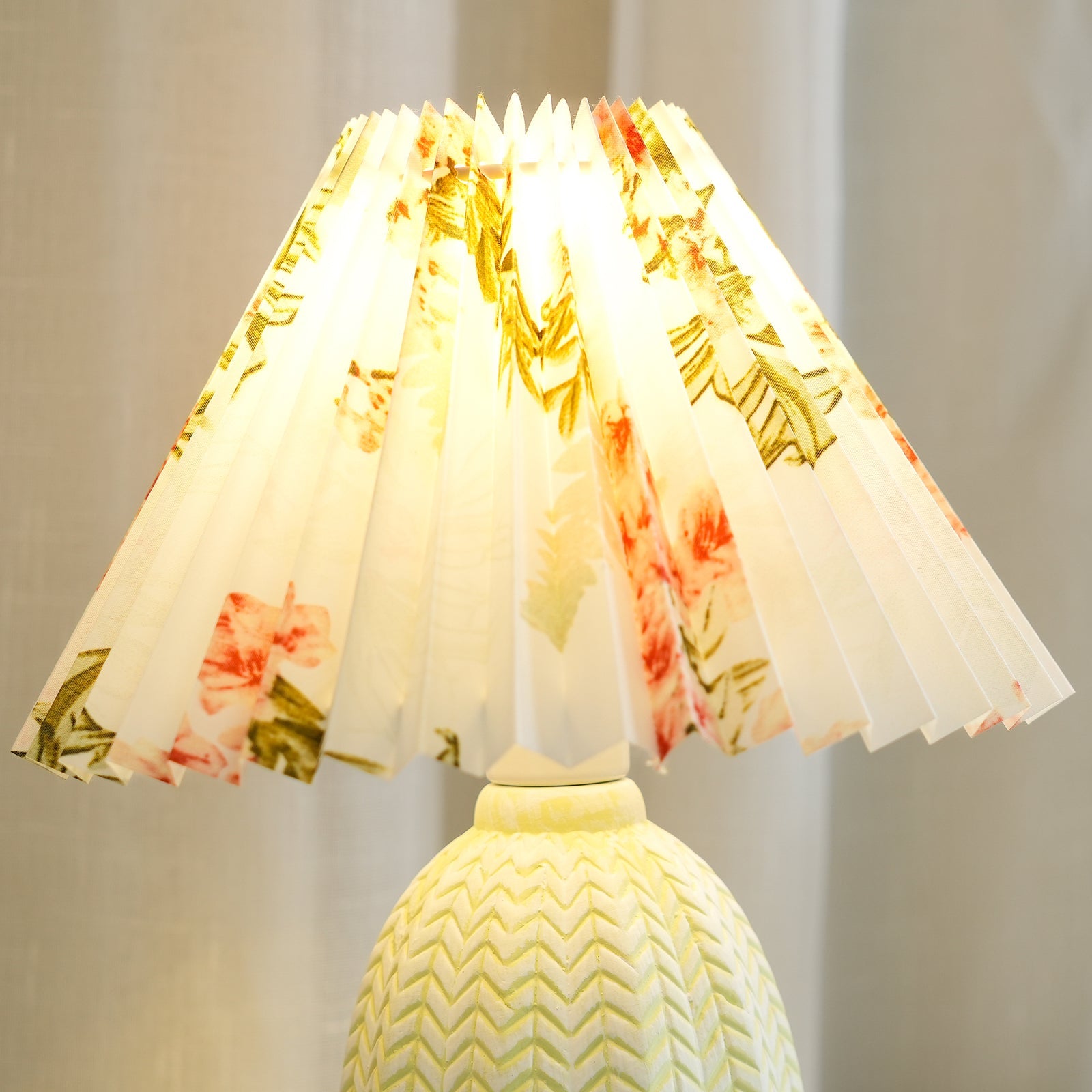 Table lamp with a white pleated shade in a printed floral pattern and a pale green ceramic base with a raised chevron pattern lit, seen close.