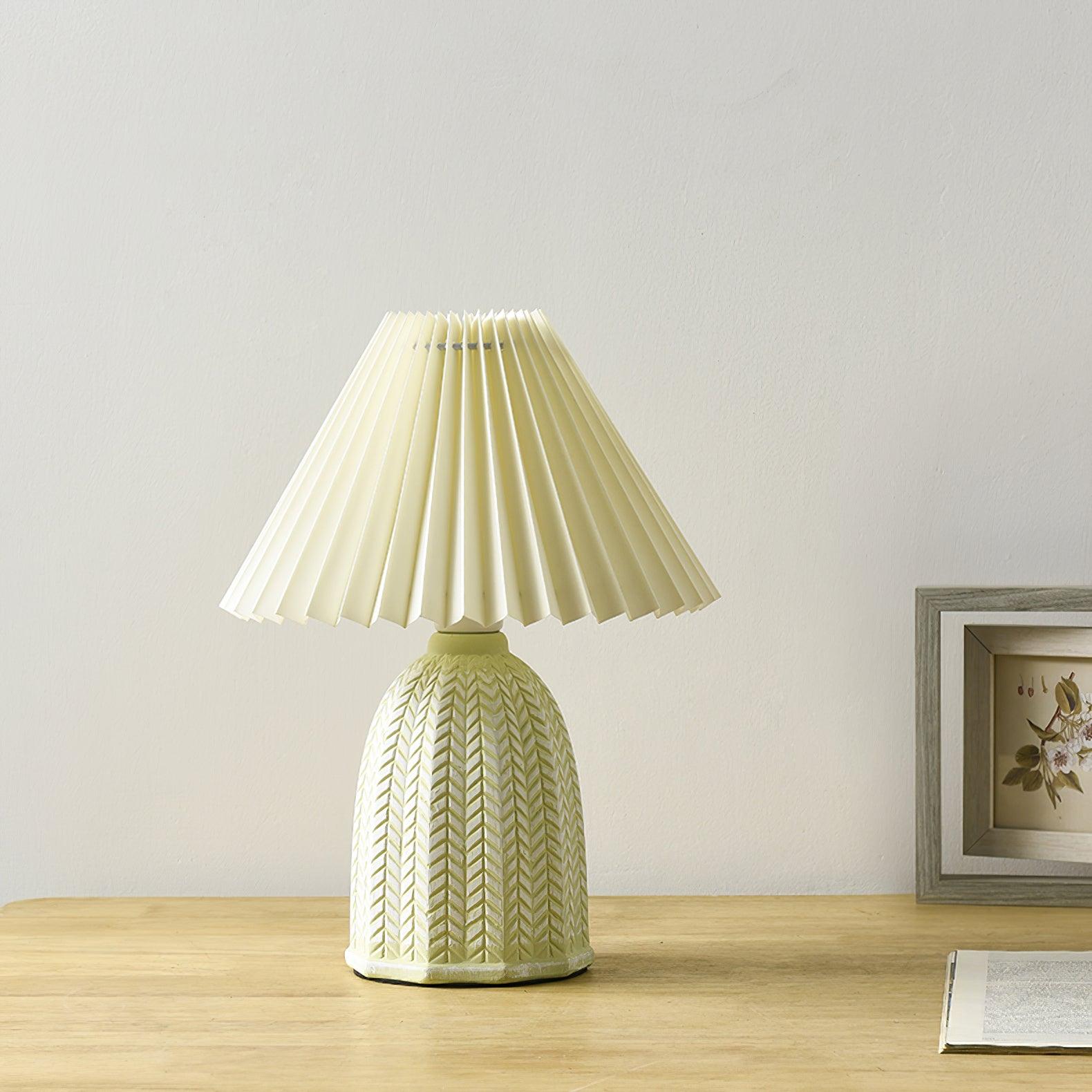 Table lamp with a plain cream pleated shade and a pale green ceramic base with a raised chevron pattern on a wooden table beside a framed picture.