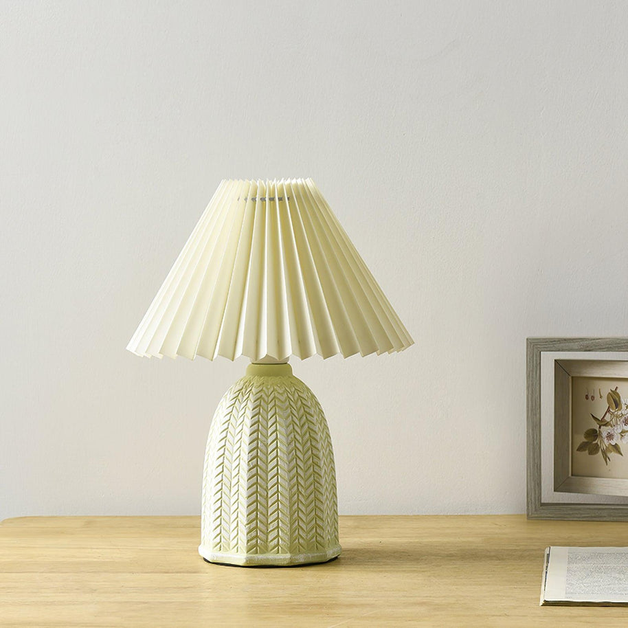 Table lamp with a plain cream pleated shade and a pale green ceramic base with a raised chevron pattern on a wooden table beside a framed picture.