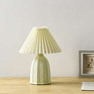 Table lamp with a plain cream pleated shade and a pale green ceramic base with a raised chevron pattern on a wooden table beside a framed picture.