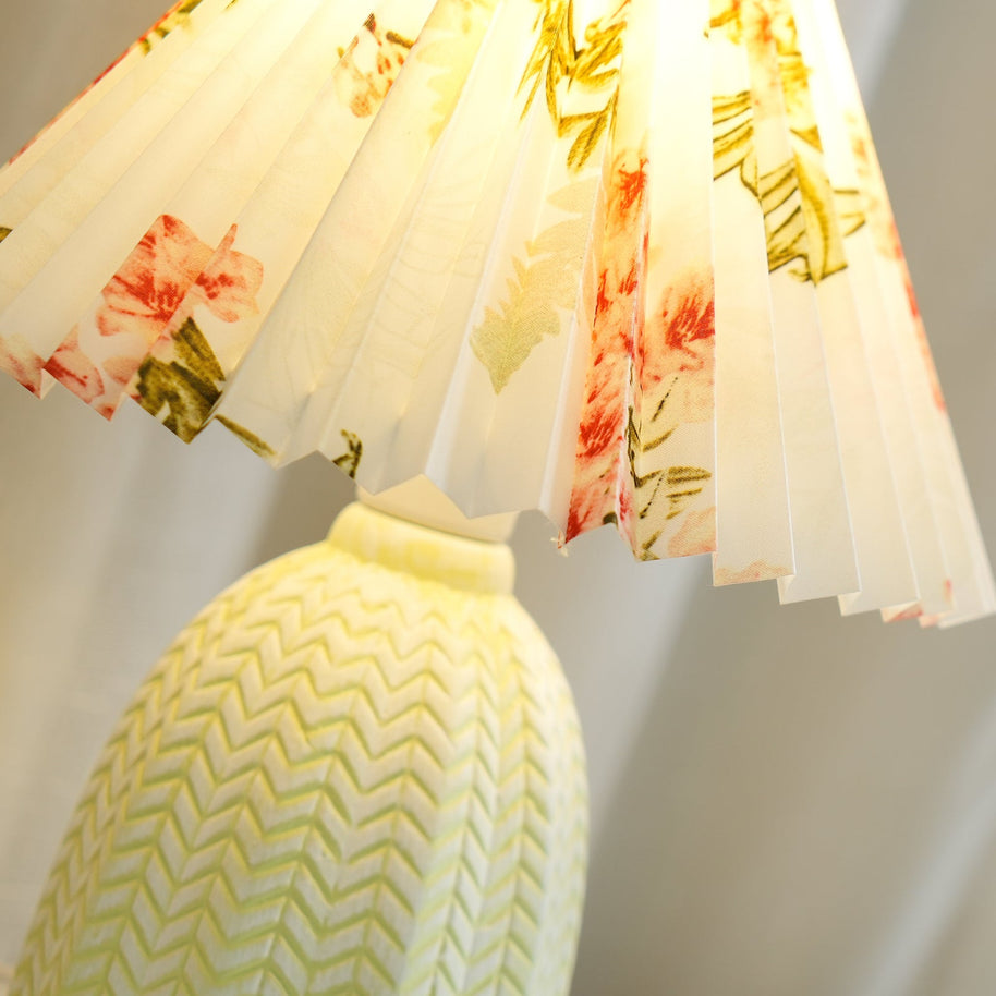 Table lamp with a white pleated shade in a printed floral pattern and a pale green ceramic base with a raised chevron pattern lit, seen close from below.