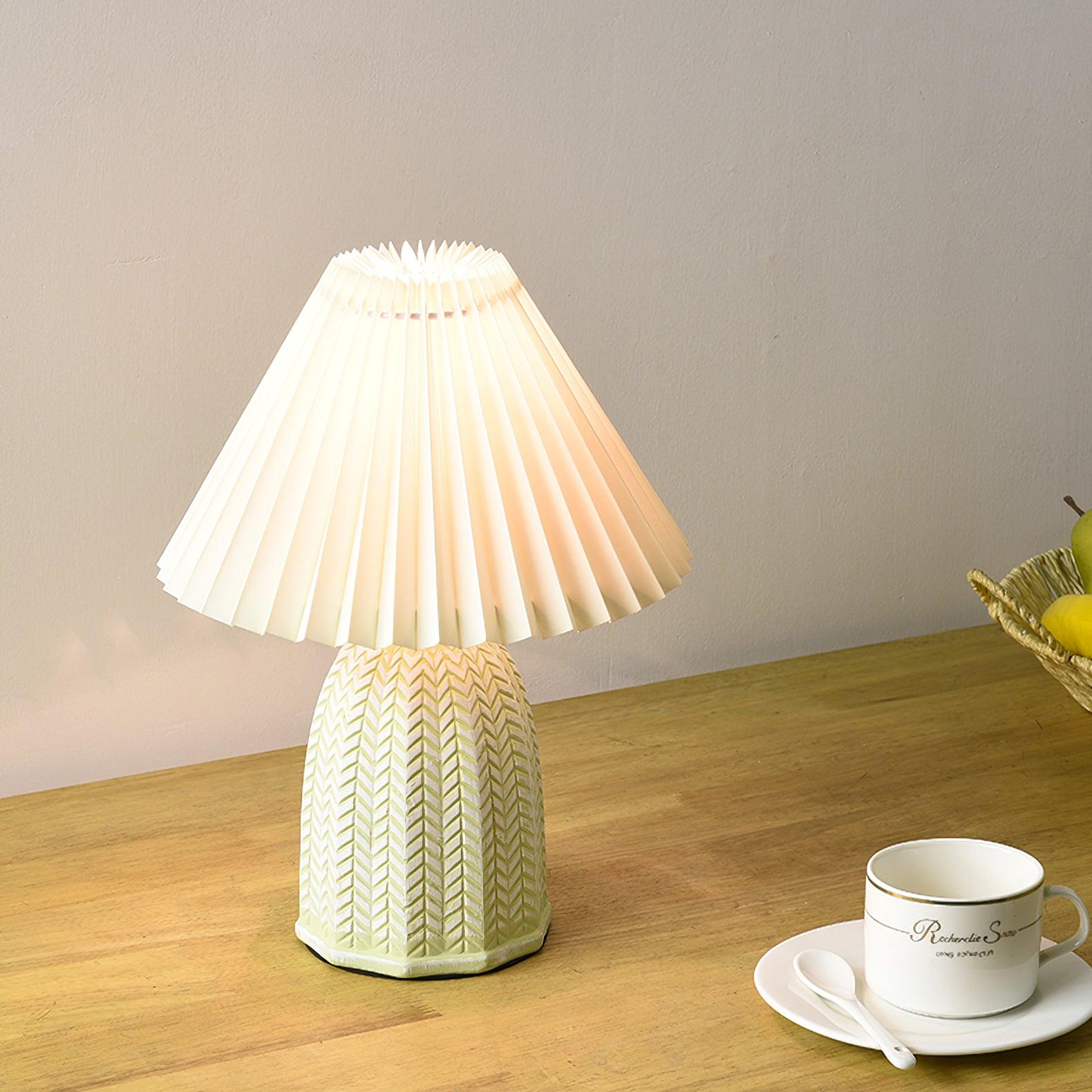 Table lamp with a plain cream pleated shade and a pale green ceramic base with a raised chevron pattern lit on a wooden table beside a teacup.