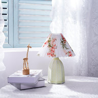 Table lamp with a white pleated shade in a printed floral pattern and a pale green ceramic base with a raised chevron pattern on a windowsill beside a book and a wooden figure.