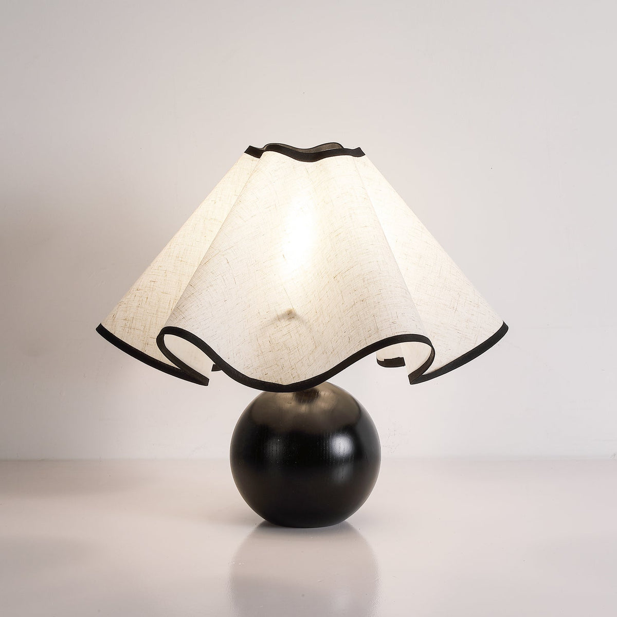 Lit table lamp with a wavy black-trimmed linen shade on a turned black sphere base, against a pale wall.
