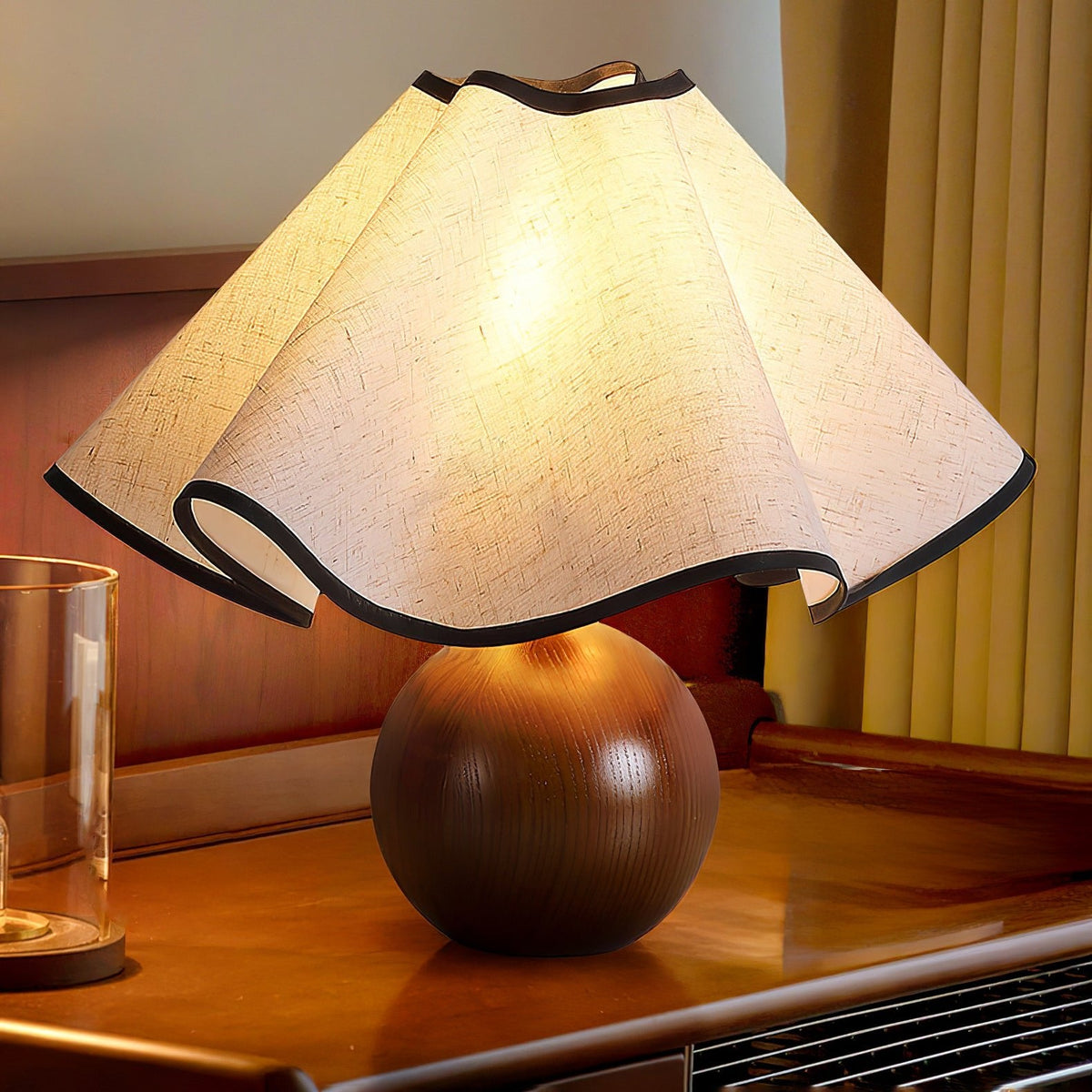 Close view of a lit table lamp: wavy black-trimmed linen shade on a walnut sphere base on a wooden desk.