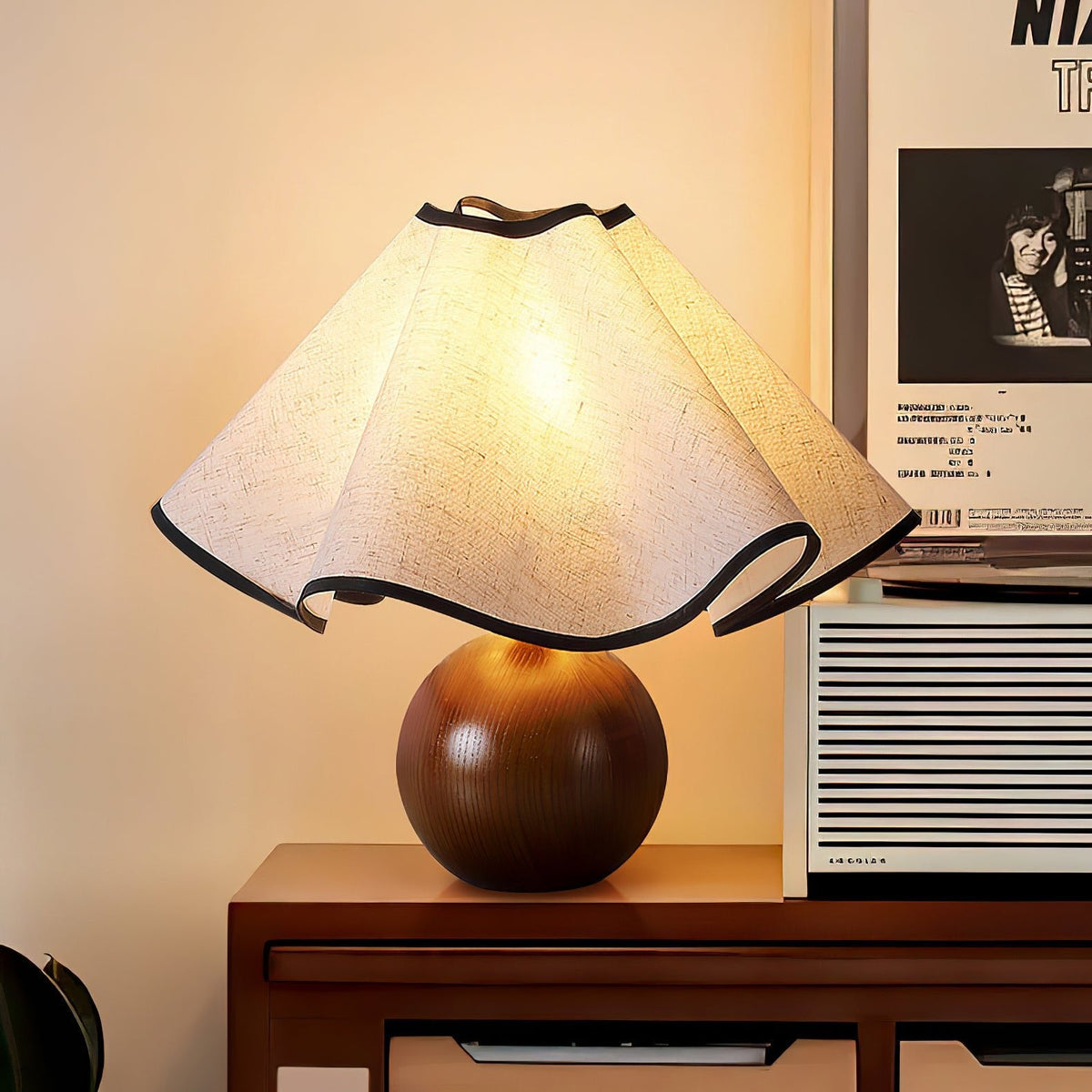 Lit table lamp with a wavy black-trimmed linen shade on a walnut sphere base on a wooden sideboard.