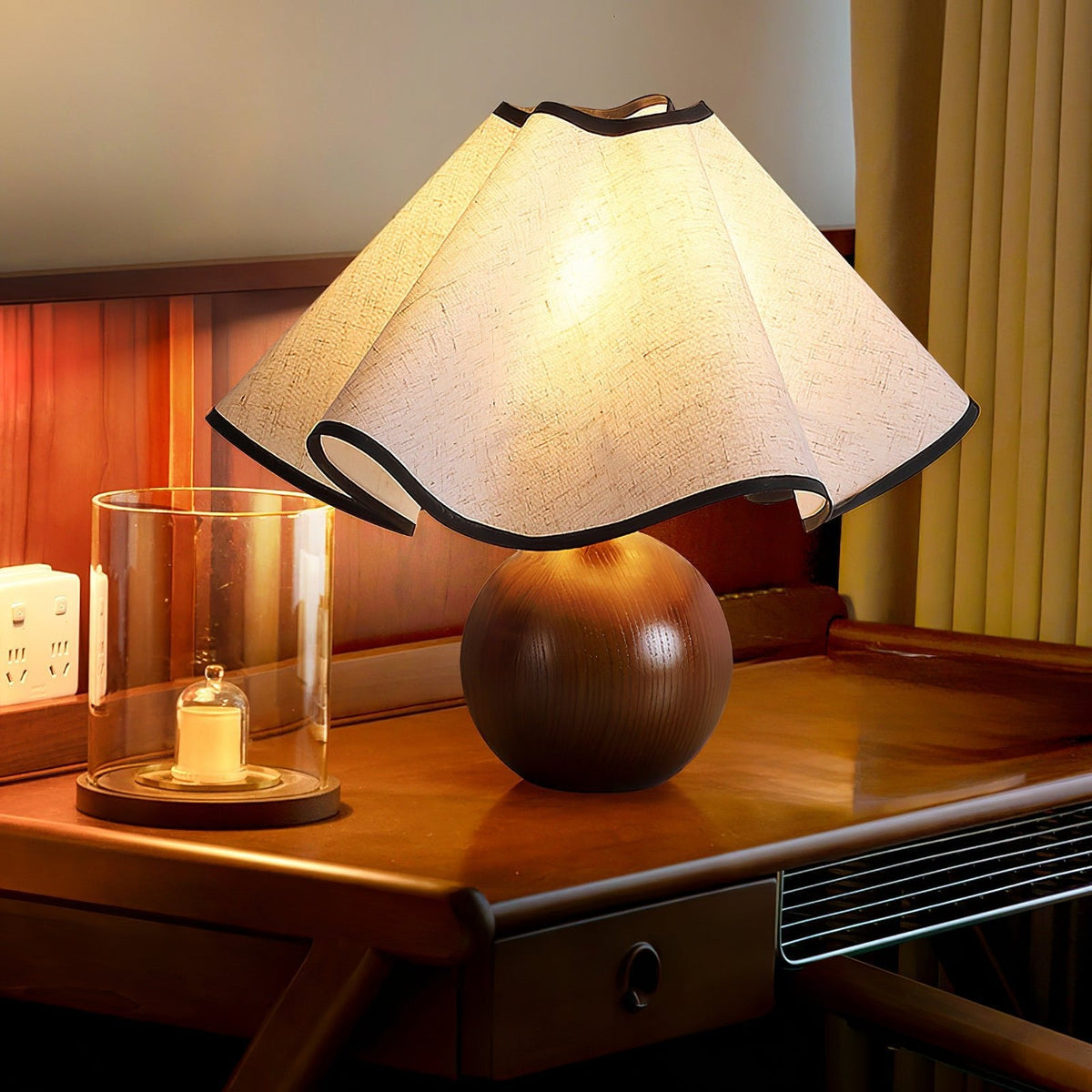 Lit table lamp with a wavy linen shade on a walnut sphere base, standing on a polished wooden desk.
