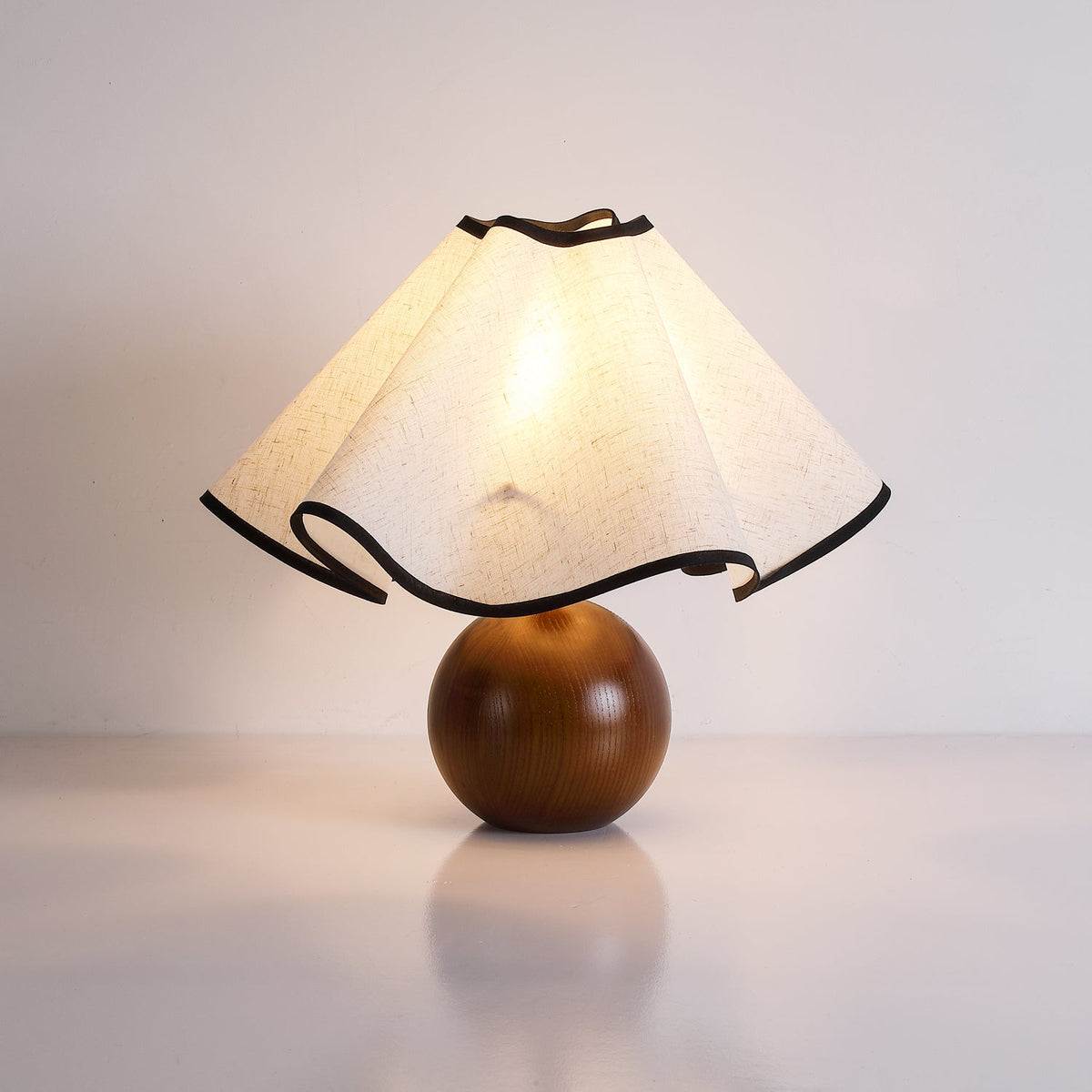 Lit table lamp with a wavy black-trimmed linen shade on a turned walnut sphere base, against a pale wall.